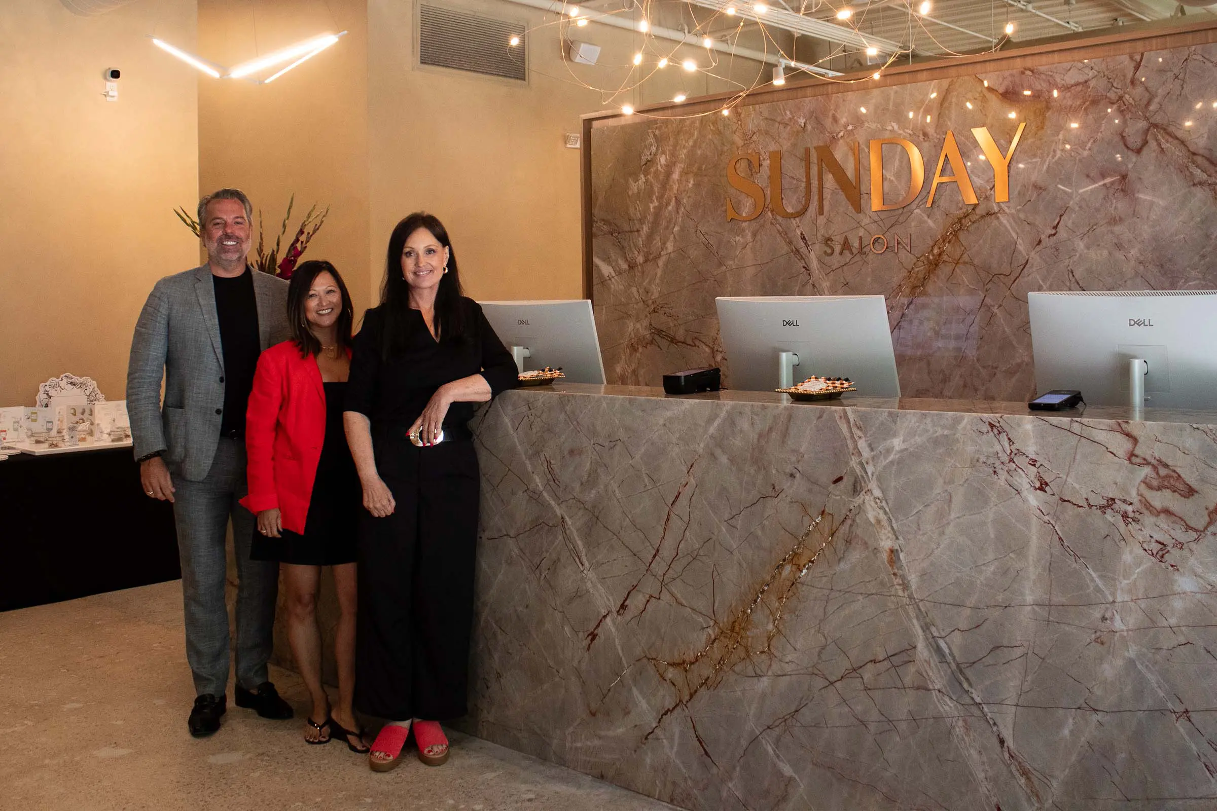 Three people stand smiling at the marble front desk of Sunday Salon, a Davines salon Raleigh, with gold letters on the wall and computer monitors on the counter. The space is modern and warmly lit with string lights above.