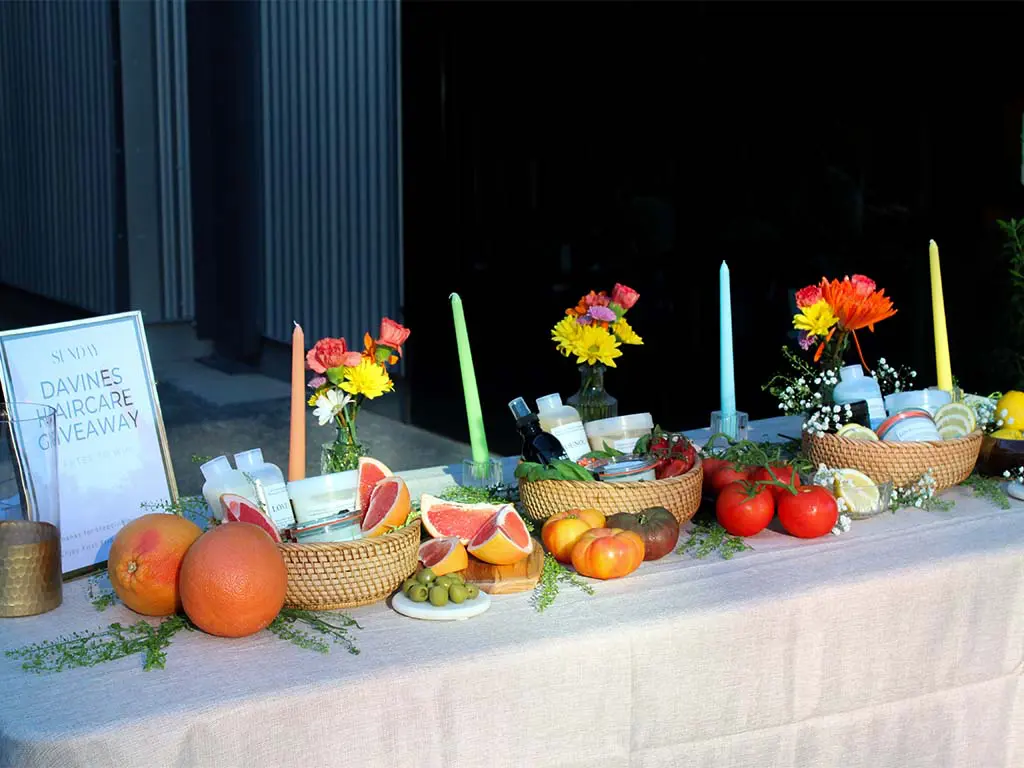 A table with assorted fruits, vegetables, flowers, candles, and haircare products. A framed sign reads “Davines Haircare Giveaway.” The setup is outdoors with a neutral tablecloth.