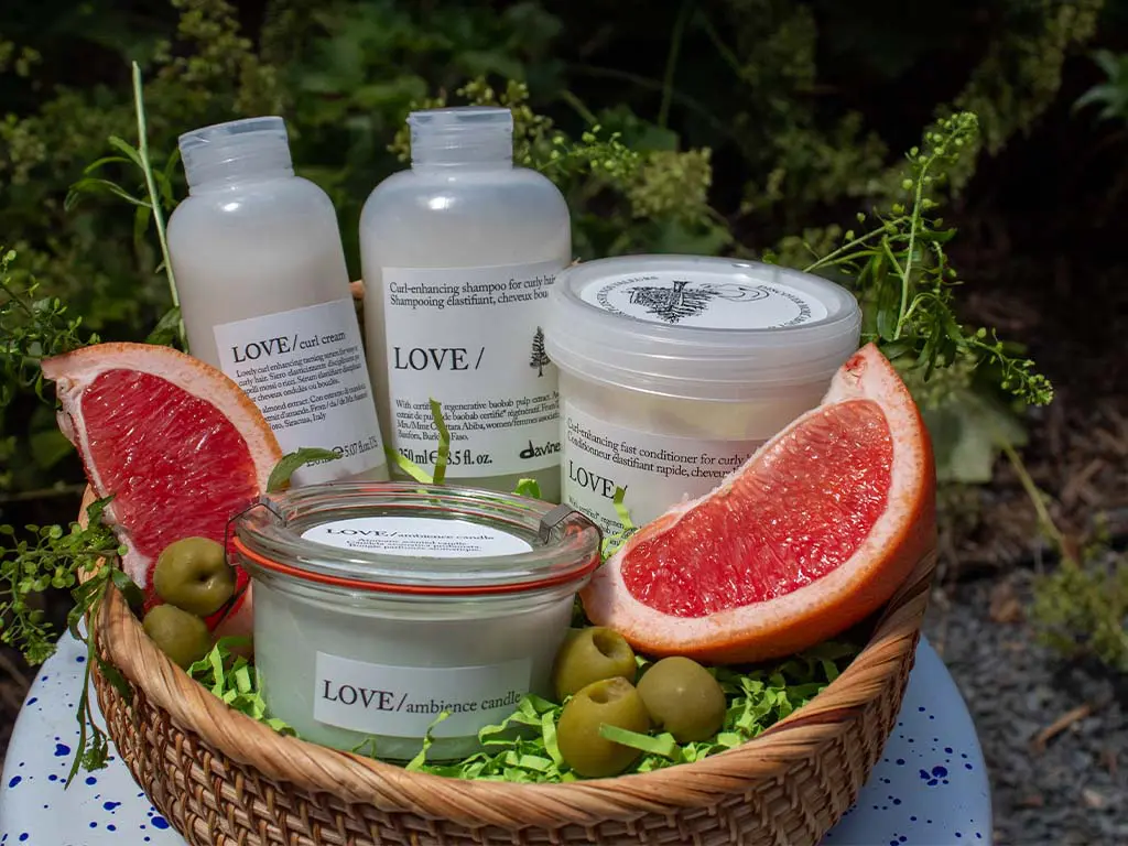 A wicker basket holds hair and skincare products, a pink grapefruit cut in half, green olives, and green shredded paper, set outdoors with greenery in the background.