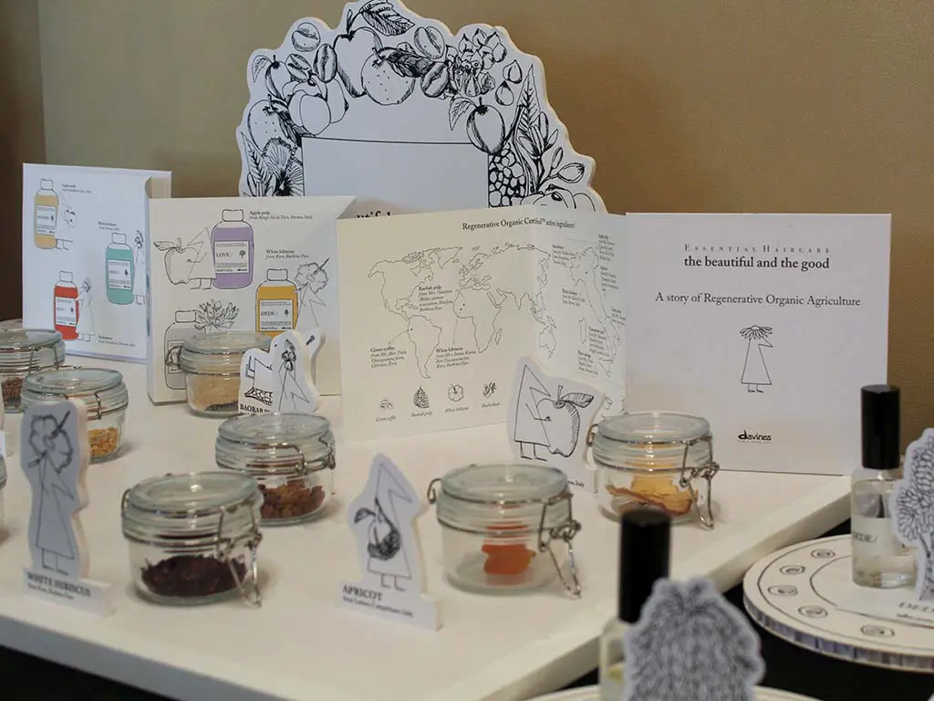 davinestable2 Display of jars with seeds and grains, labeled with crop illustrations and names like wheat and apricot, alongside informational cards about regenerative organic agriculture on a white table.