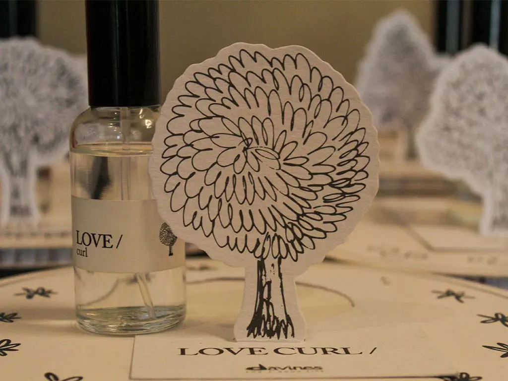 davinestable3 A small bottle labeled “LOVE curl” stands next to a paper cut-out of a tree with curly branches, all placed on a surface decorated with similar tree illustrations.