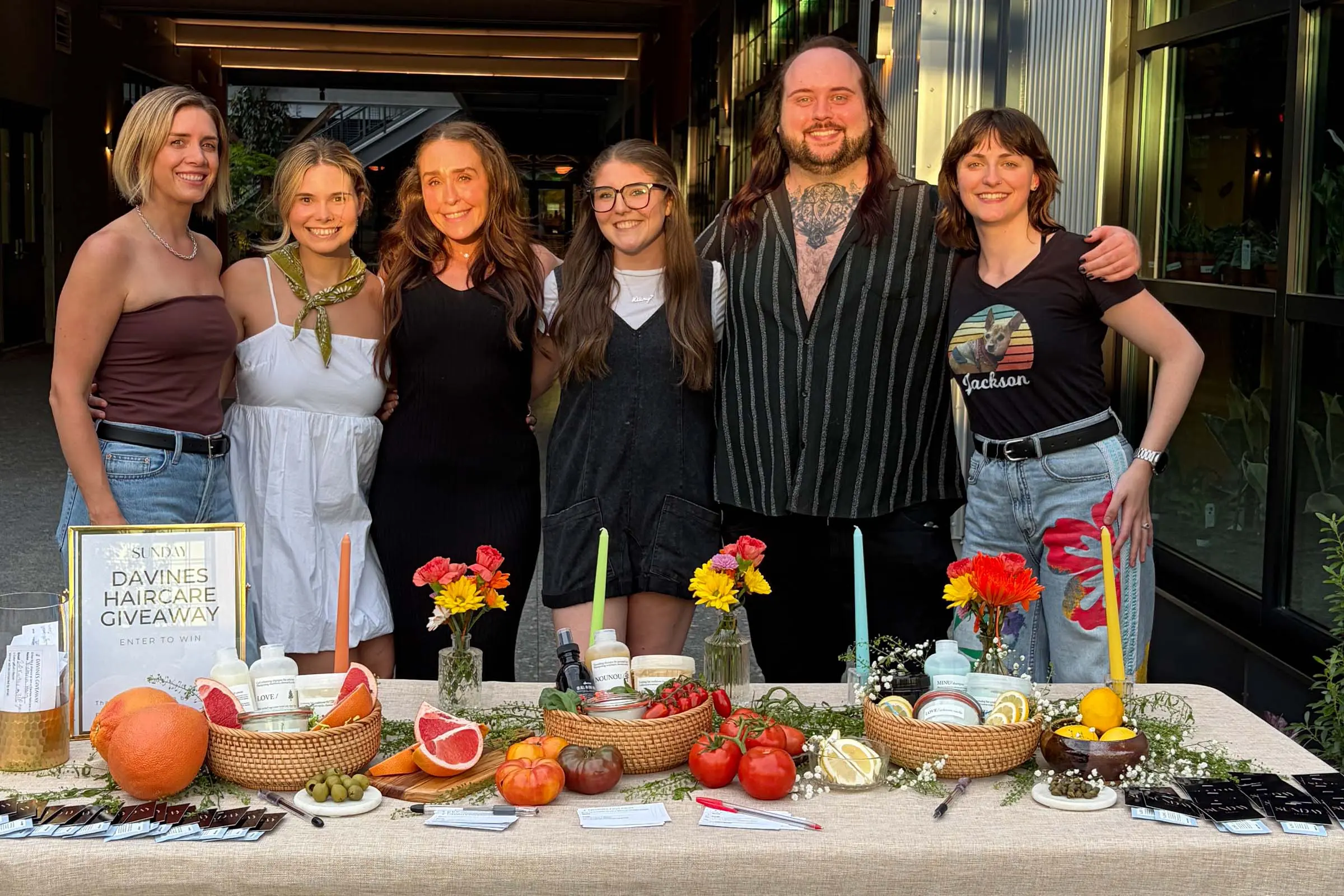 Six people smile behind a table with fruit, flowers, candles, and haircare products on display. A sign reads “Davines Haircare Giveaway.” They stand outside a modern building in natural light, representing a Raleigh hair salon committed to beauty and community.