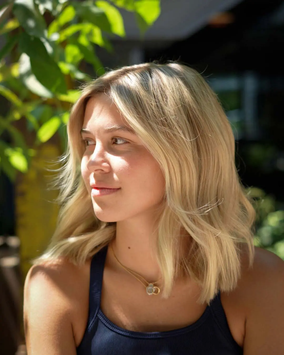 nathan-jolly-lived-in-blonde-haircut-sunday-salon-raleigh A young woman with shoulder-length lived-in-blonde hair looks to the side in sunlight, wearing a sleeveless navy top and a gold necklace, with green leaves and a blurred outdoor background behind her—embodying the effortless style often seen at Sunday Salon Raleigh.