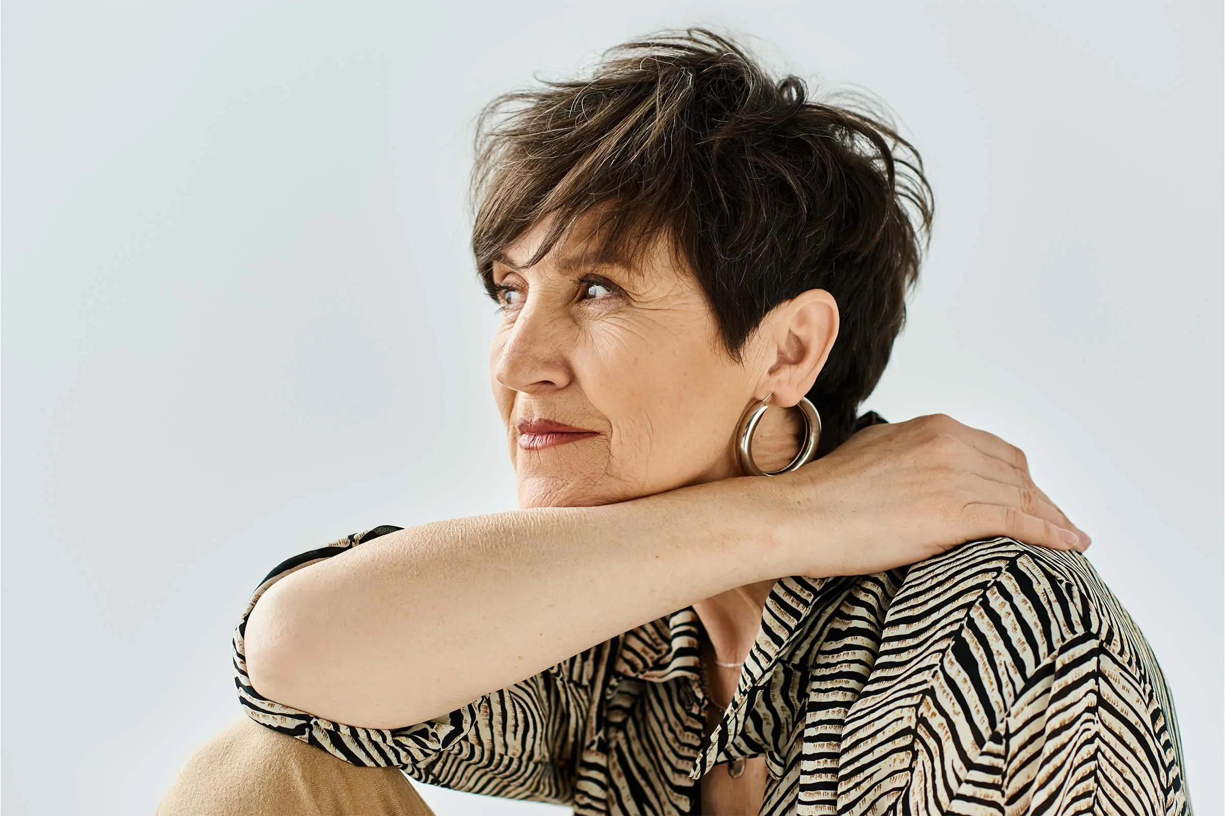 A woman with short brown precision haircut and hoop earrings gazes thoughtfully to the side, resting her hand on her shoulder and wearing a striped shirt against a plain background.