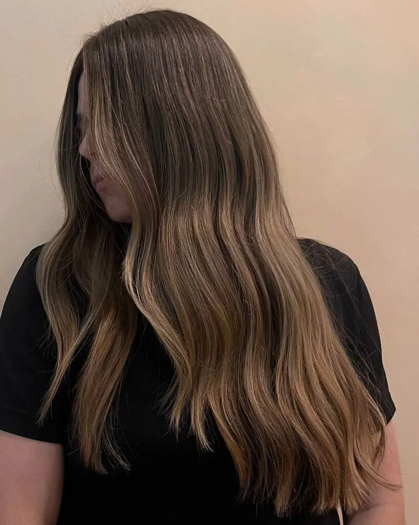 Woman with long, soft waves of warm brunette hair featuring subtle blonde highlights in a beautiful brunette balayage, wearing a black shirt and facing sideways against a plain beige background.