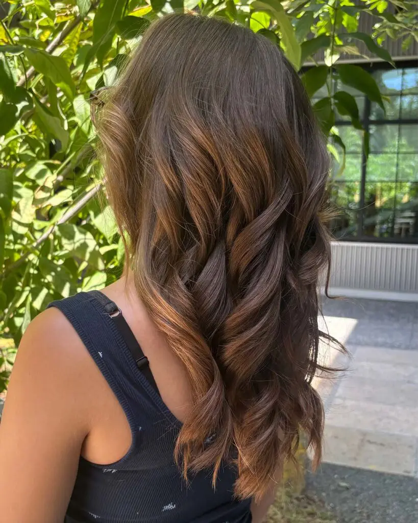 A side-back view of a woman featuring long, cascading rich chestnut waves with warm cinnamon dimension, expertly styled at Sunday Salon in Raleigh.