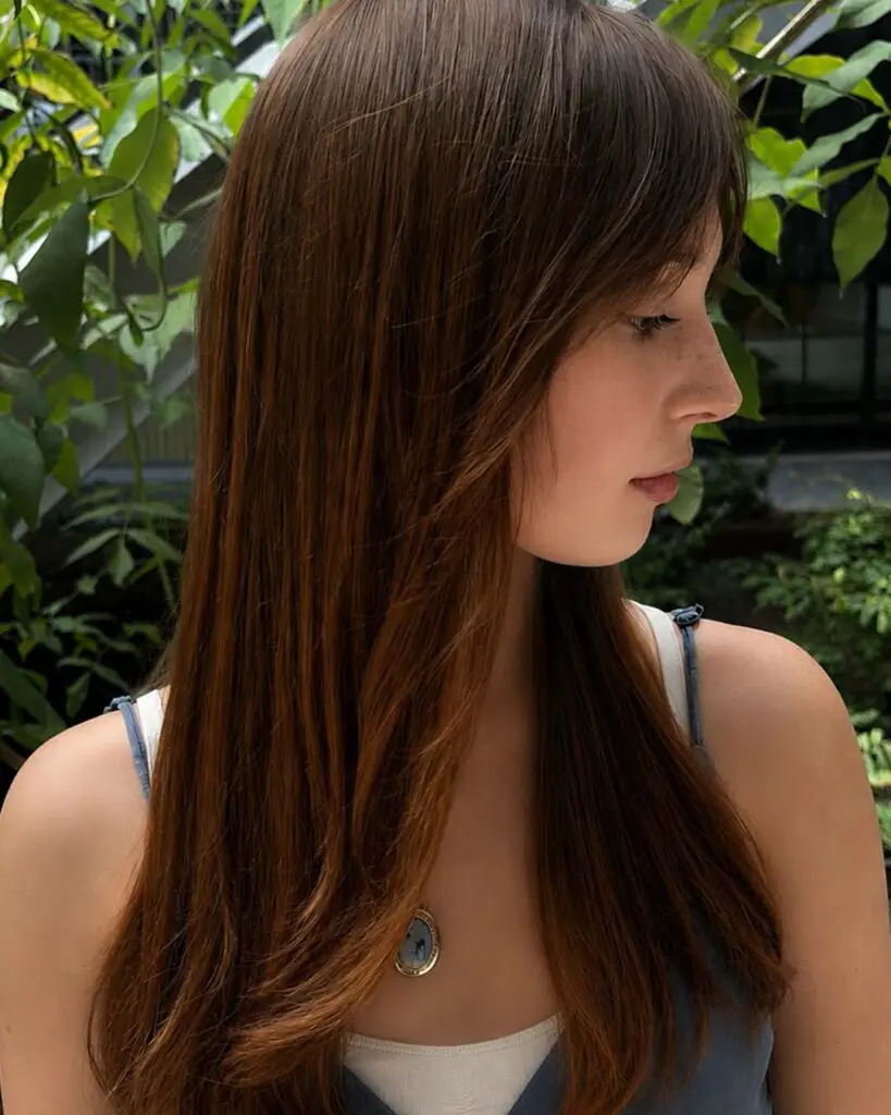 A side profile of a woman showcasing long, silky chestnut brown hair with soft curtain bangs styled at Sunday Salon in Raleigh.