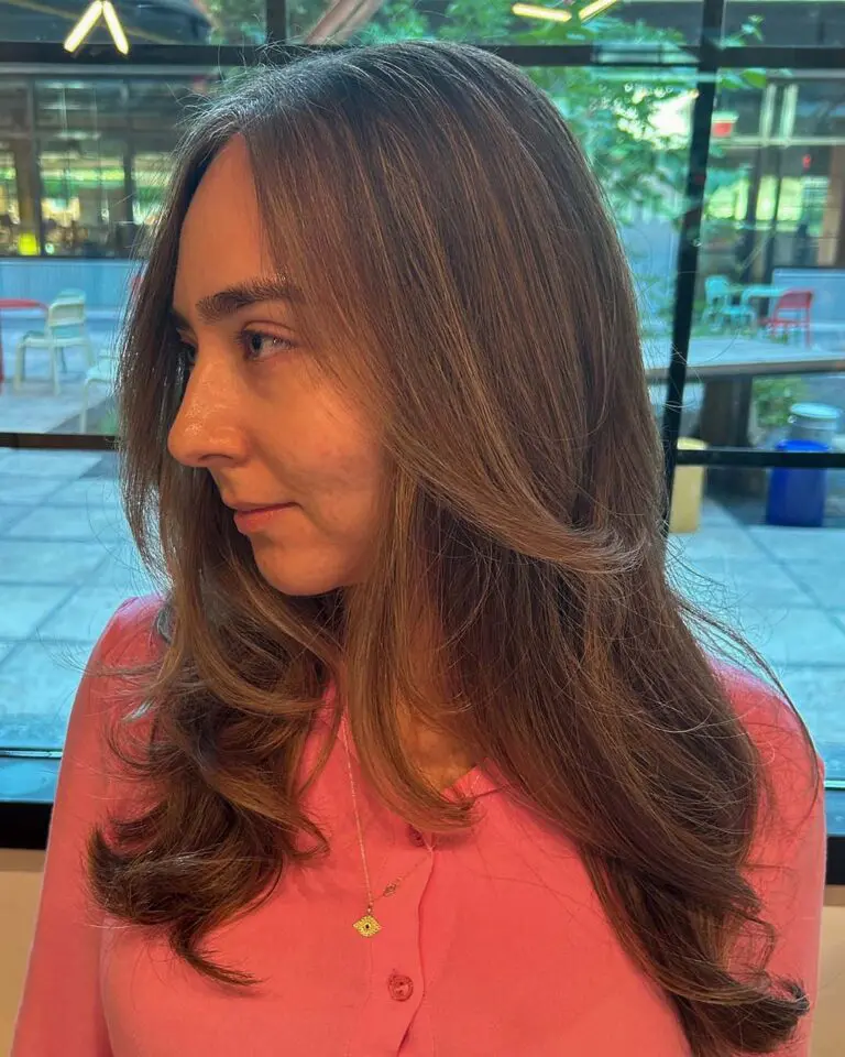 A side view of a woman featuring long, soft chestnut waves with seamless highlights and face-framing curtain bangs styled at Sunday Salon in Raleigh.