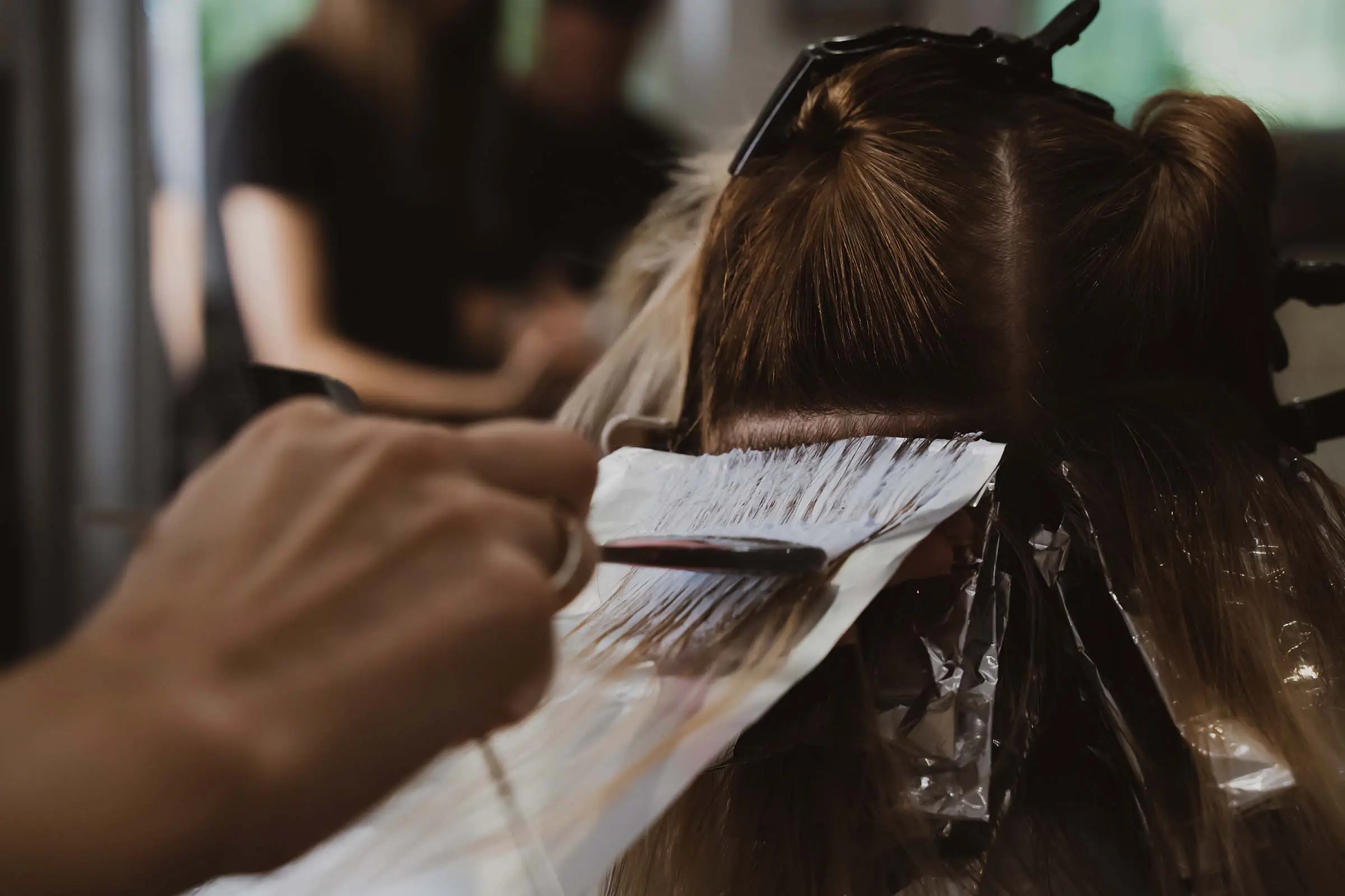 A close-up of a person’s hair being dyed at a salon, showcasing professional hair coloring services. Sections of hair are separated with foil while a stylist applies hair dye with a brush. The background is blurred.
