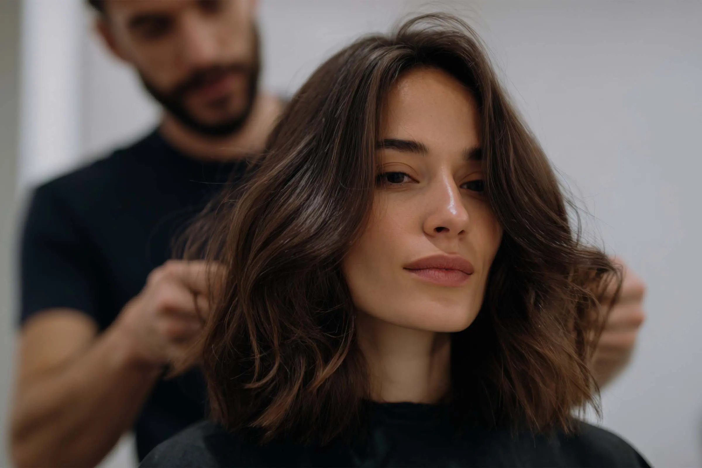 A woman with shoulder-length brown hair sits while a hairdresser styles her hair, offering a personalized hair consultation. The background is blurred, focusing on her calm expression and the hairstylist’s hands expertly arranging her hair.