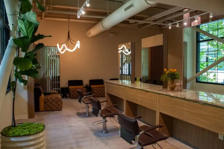 Modern hair salon Raleigh with brown chairs, mirrors, a marble counter, plants, and warm lighting. Large windows bring in natural light, creating an inviting and stylish atmosphere.