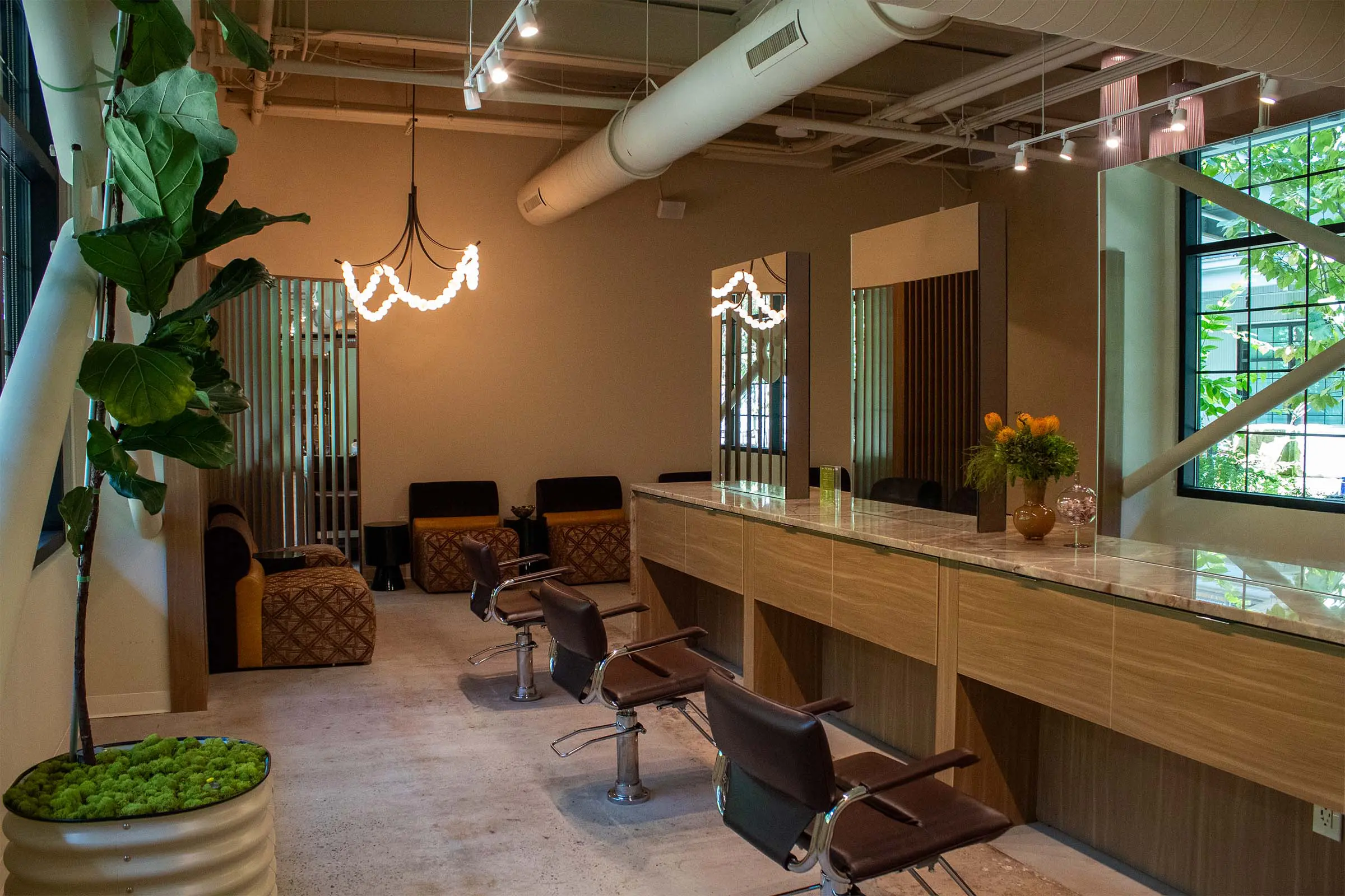 Modern hair salon Raleigh with brown chairs, mirrors, a marble counter, plants, and warm lighting. Large windows bring in natural light, creating an inviting and stylish atmosphere.