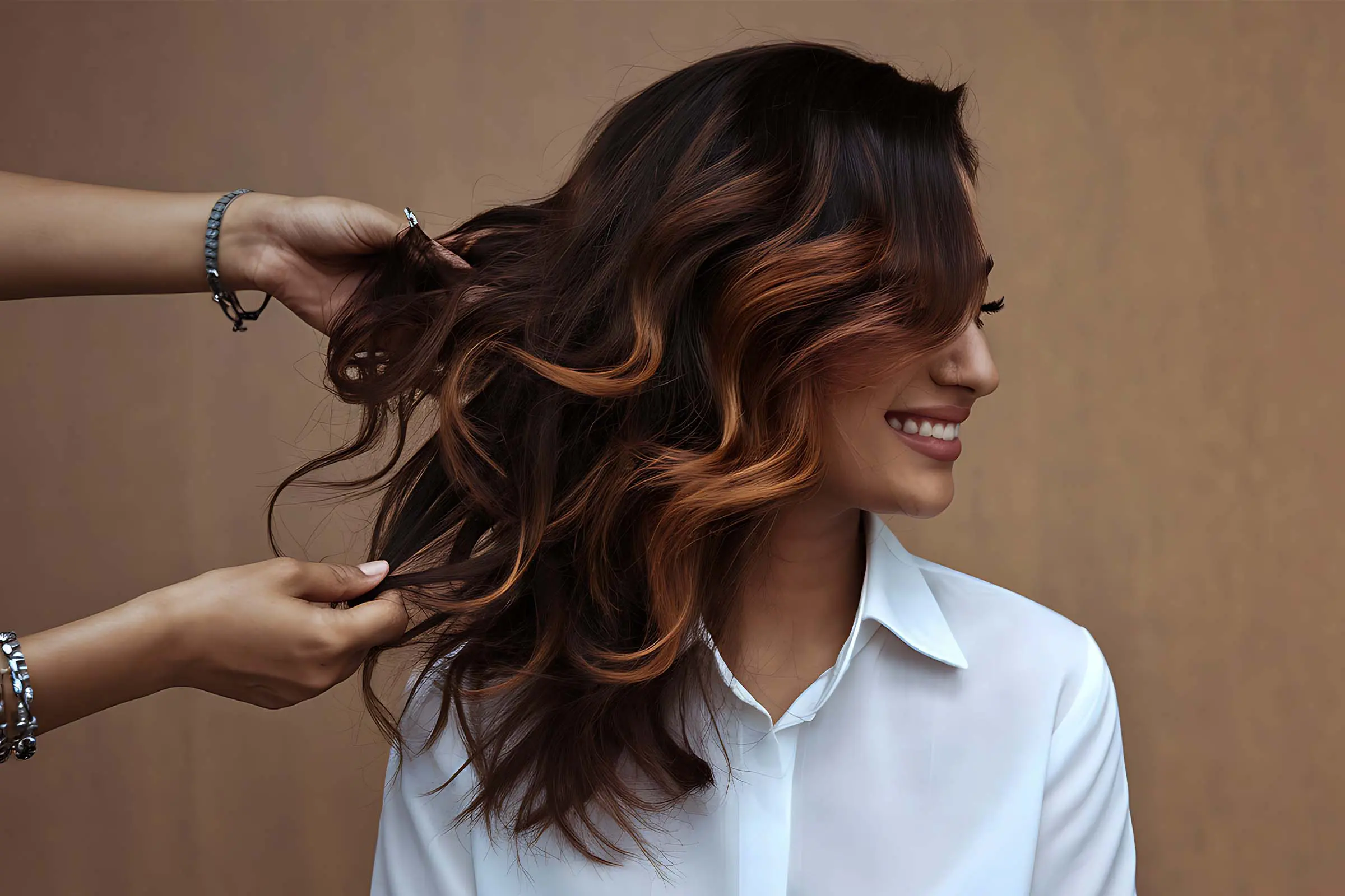 A smiling woman in a white shirt has her long, wavy, brown hair with copper highlights styled by another person against a plain tan background, perfectly capturing the essence of a seasonal hair refresh.