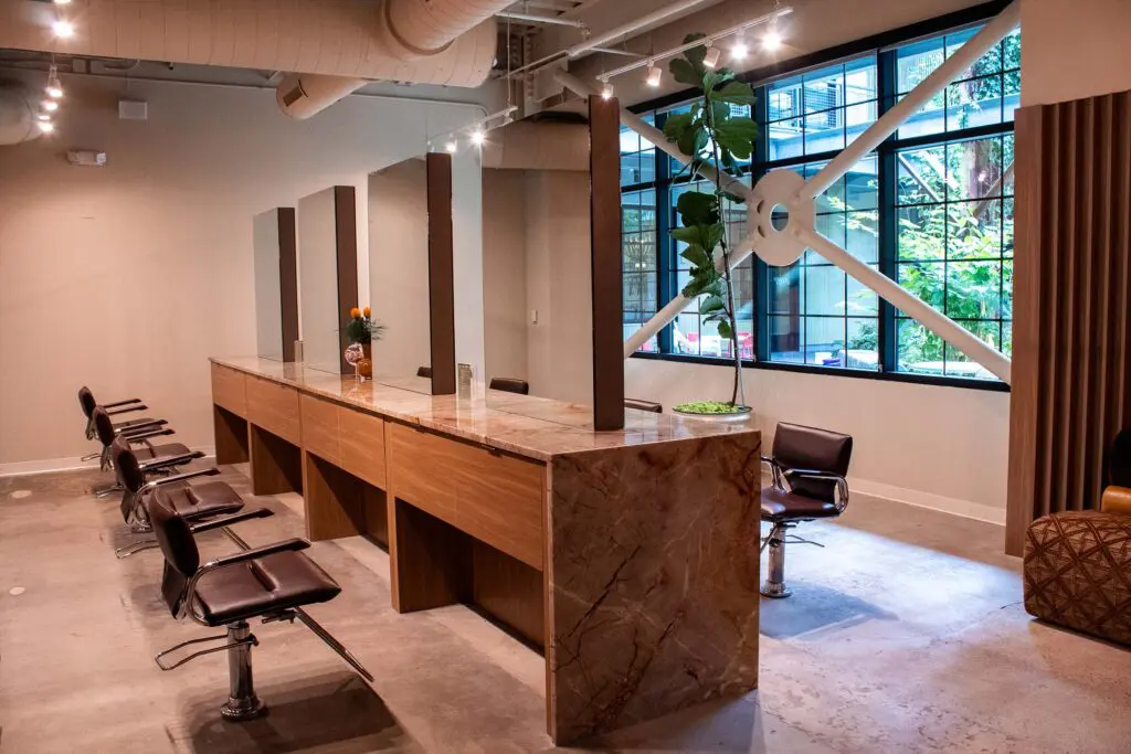 A Raleigh hair salon interior featuring a row of styling chairs and stations with marble countertops, partitioned by mirrors and tall windows overlooking greenery.