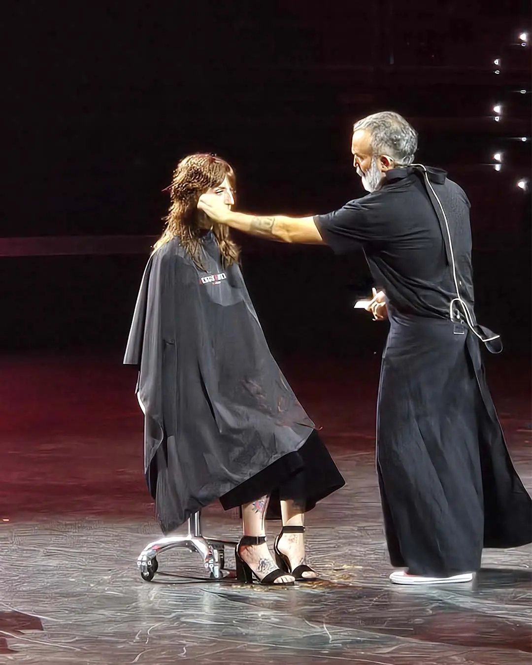davines-europe-11 A hairstylist touches the face of a client sitting in a salon chair on a stage.