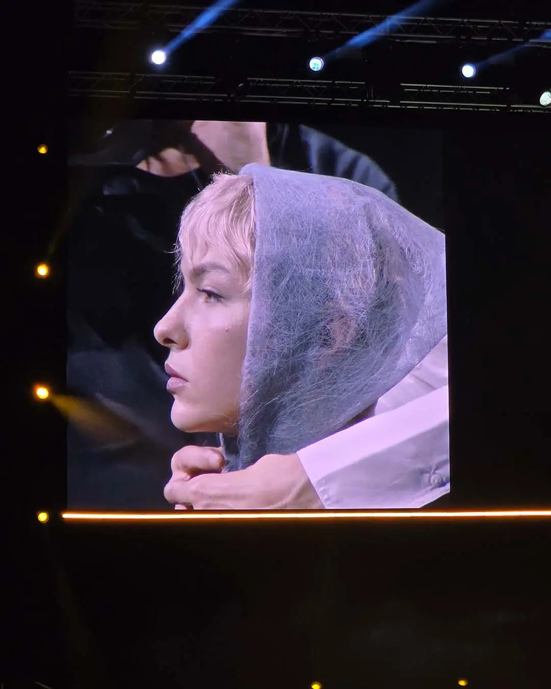 davines-europe-3 A person with short blonde hair wears a light blue translucent veil draped over their head, shown on a large concert screen in profile.