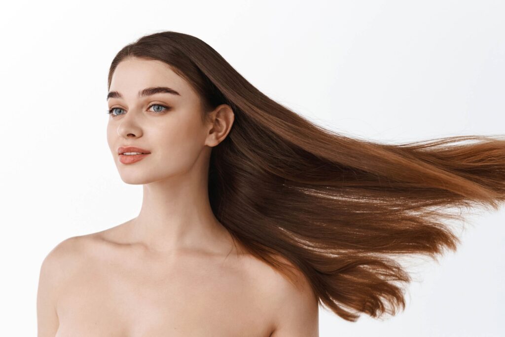 A woman with long, straight brown hair is shown from the shoulders up against a white background. Her hair, styled beautifully as if by a professional hair salon, is flowing to the side, and she has a serene expression on her face. She has light skin and blue eyes and is looking slightly away from the camera.