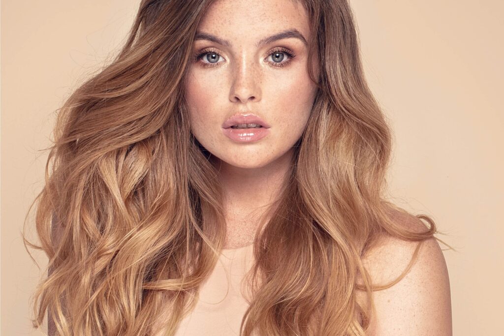 A woman with wavy, shoulder-length blonde hair and light complexion looks directly at the camera. She has a neutral expression, light makeup, glossy lips, and is wearing a beige top. The background is a plain, matching beige color—perfectly styled by the best hair salon Cary NC has to offer.