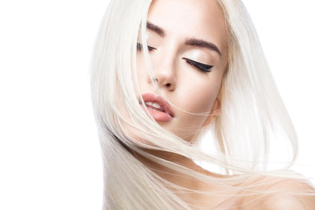A person with long, flowing platinum blonde hair is seen with closed eyes and slightly parted lips. Their makeup includes winged eyeliner and a light, shimmery eyeshadow. The background is plain white, emphasizing the person's serene and elegant expression—perhaps captured at the best hair salon Cary NC has to offer.
