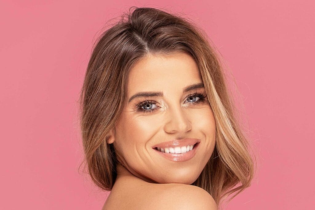 A woman with light brown hair accented by a partial balayage smiles confidently at the camera. Her bright blue eyes and soft pink lipstick complement her natural makeup look. She is posed against a solid pink background, radiating charm in Cary, NC.