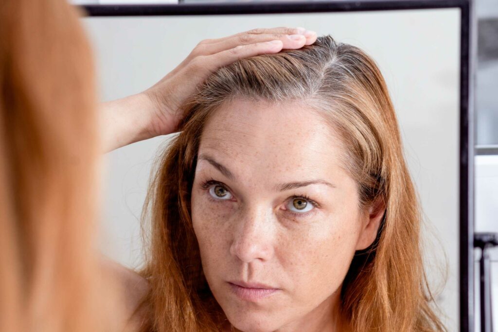 A woman with light brown hair is looking into a mirror, parting her hair to closely inspect the roots. She has a concerned expression on her face as she examines some visible grey strands, considering whether it's time for a visit to her favorite hair salon. The background is out of focus, highlighting her reflection.