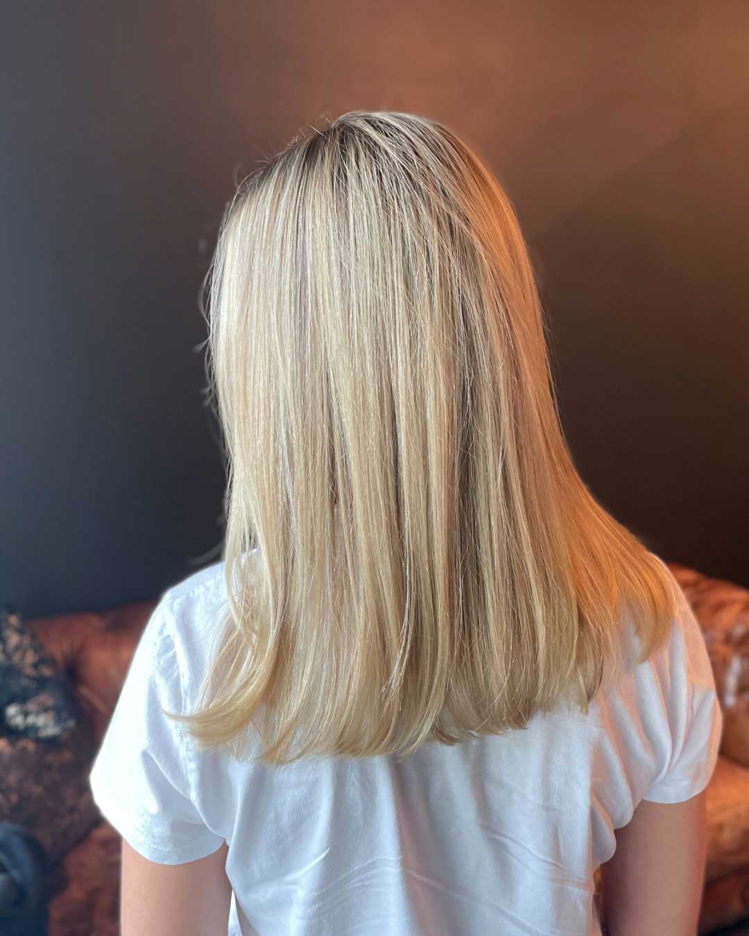 A person with straight blonde hair, wearing a white shirt, is facing away from the camera. The background includes a dark-colored wall and couch.