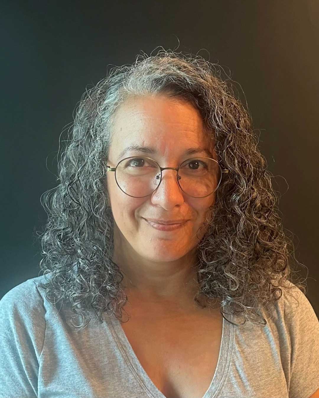 A woman with curly salt-and-pepper hair and wearing glasses smiles softly at the camera. She is dressed in a light gray shirt and stands against a dark background.