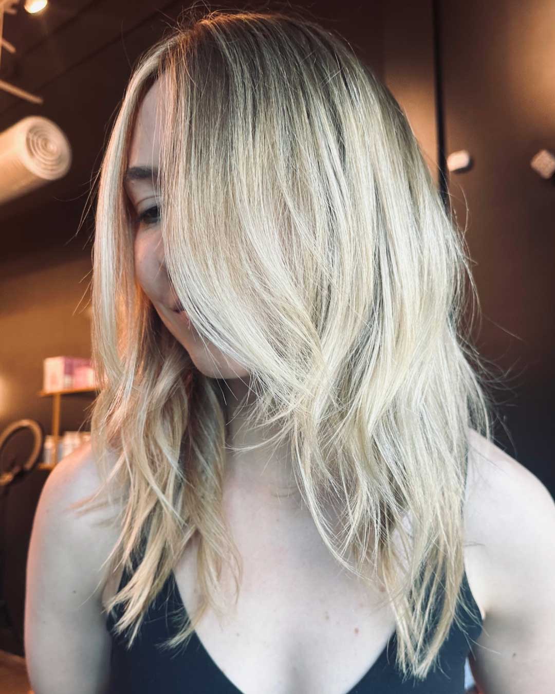 A woman with light blonde hair styled in loose waves stands against a dark indoor background. She is wearing a black top, and her long hair partially covers her face, revealing a subtle smile.