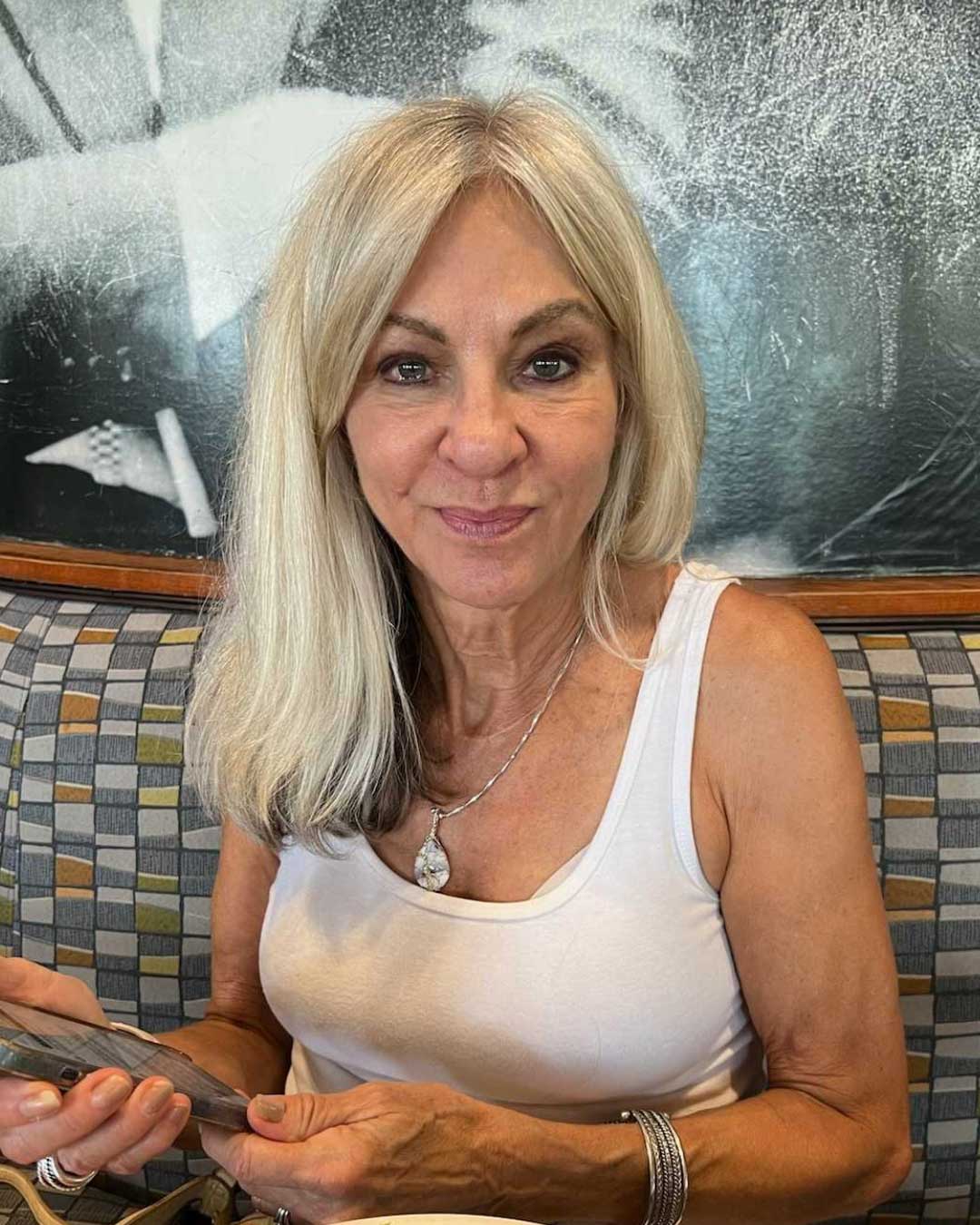 A woman with long, silver hair sits on a cushioned bench. She is wearing a white tank top and several bracelets, holding a cellphone. The background is a textured, abstract artwork. She is smiling softly and looking directly at the camera.