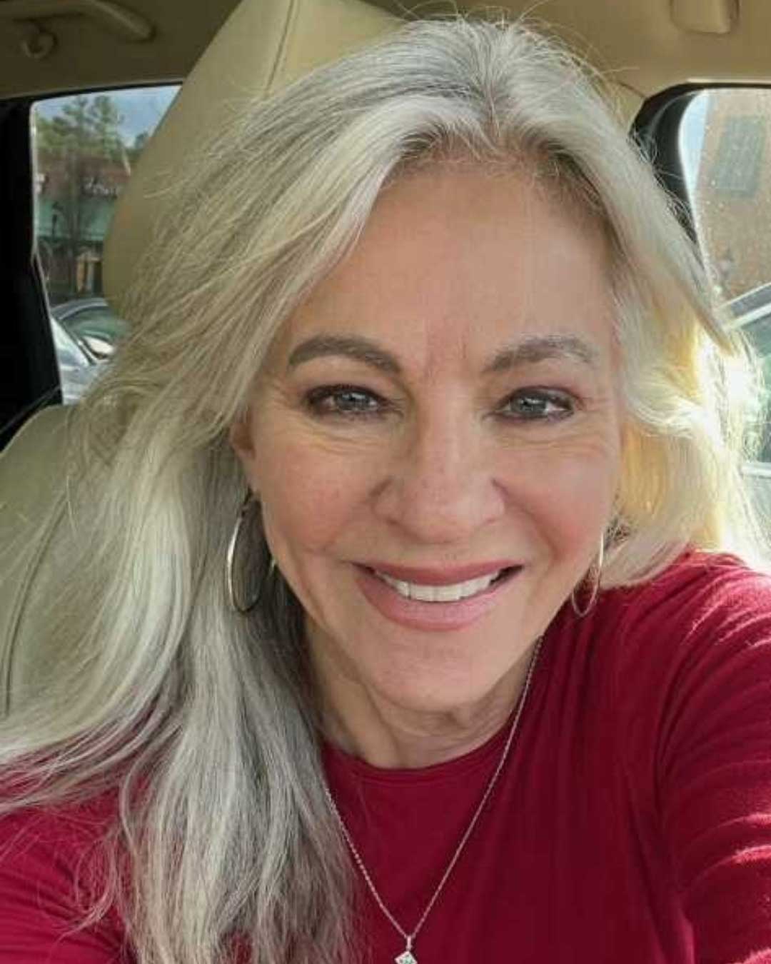 A woman with long, wavy gray hair and wearing a red top smiles at the camera. She is seated in a car with a beige interior. She has hoop earrings and is bathed in natural light.