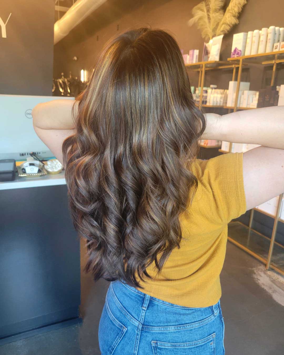 A person with long, wavy, light brown hair is shown from the back, standing in a salon. They are wearing a mustard yellow top and blue jeans, with both hands running through their hair. Shelves with hair products are visible in the background.