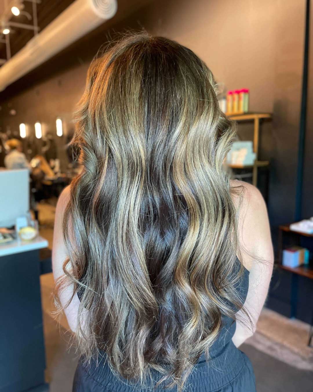 A person with long, wavy hair featuring a mix of blonde and brown highlights, standing indoors in what appears to be a salon. The background shows shelves with various hair care products and a modern, stylish decor. The person's face is not visible.