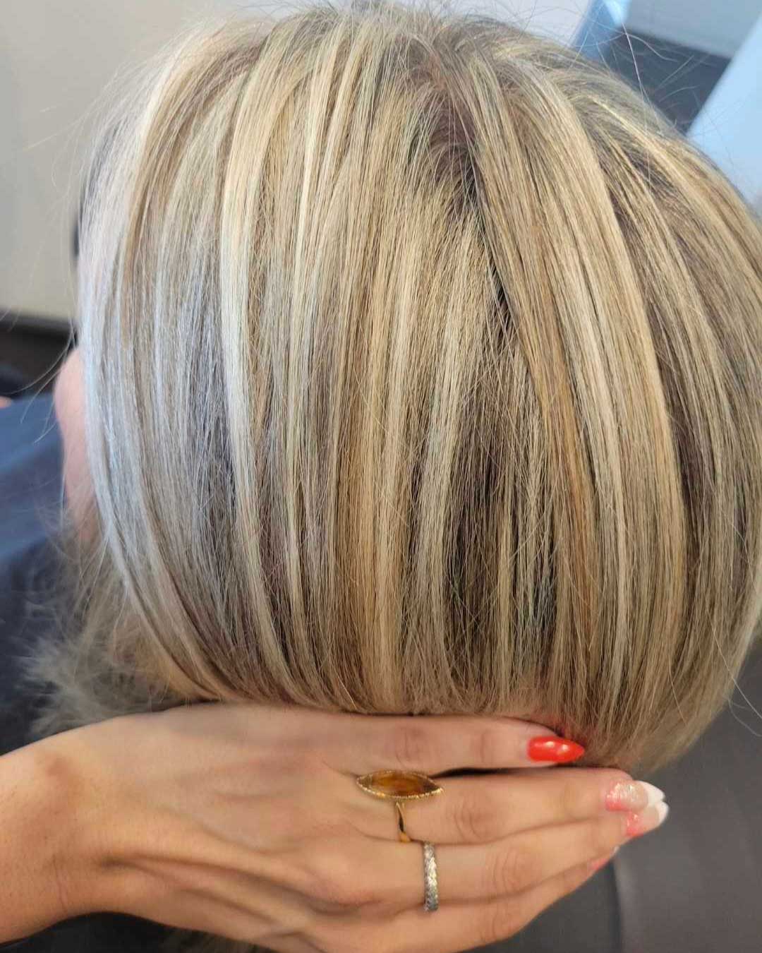 A close-up of a person's head showing freshly highlighted blonde hair with darker roots. The hair is styled straight and glossy. The person’s hand with red-painted nails and a ring is gently touching the back of their head.