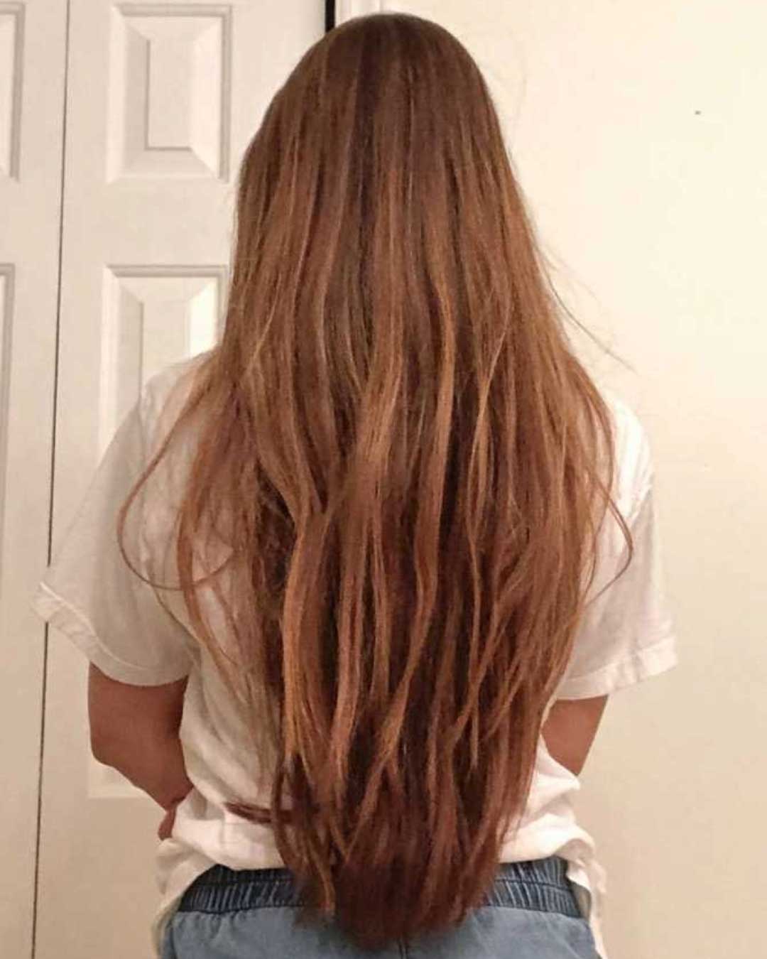 A person with long brown hair, seen from behind, stands in front of a beige wall and white door. They are wearing a white shirt and blue jeans. The hair reaches down to their waist, flowing naturally without any visible styling.