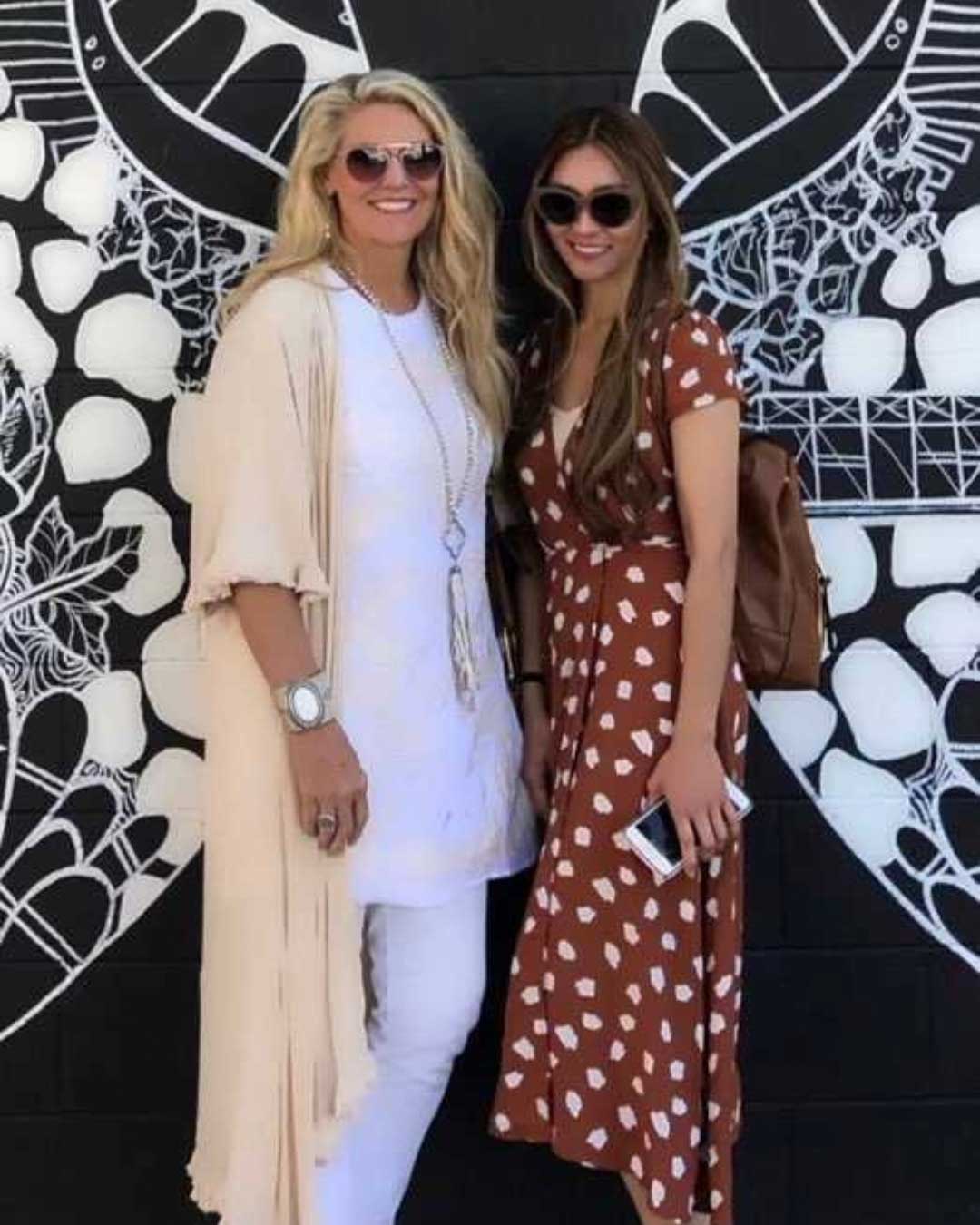 Two women stand in front of a black wall adorned with white, intricate, wing-like artwork. Both are wearing sunglasses and smiling. The woman on the left wears a long beige cardigan over a white outfit, while the woman on the right wears a brown dress with white spots.