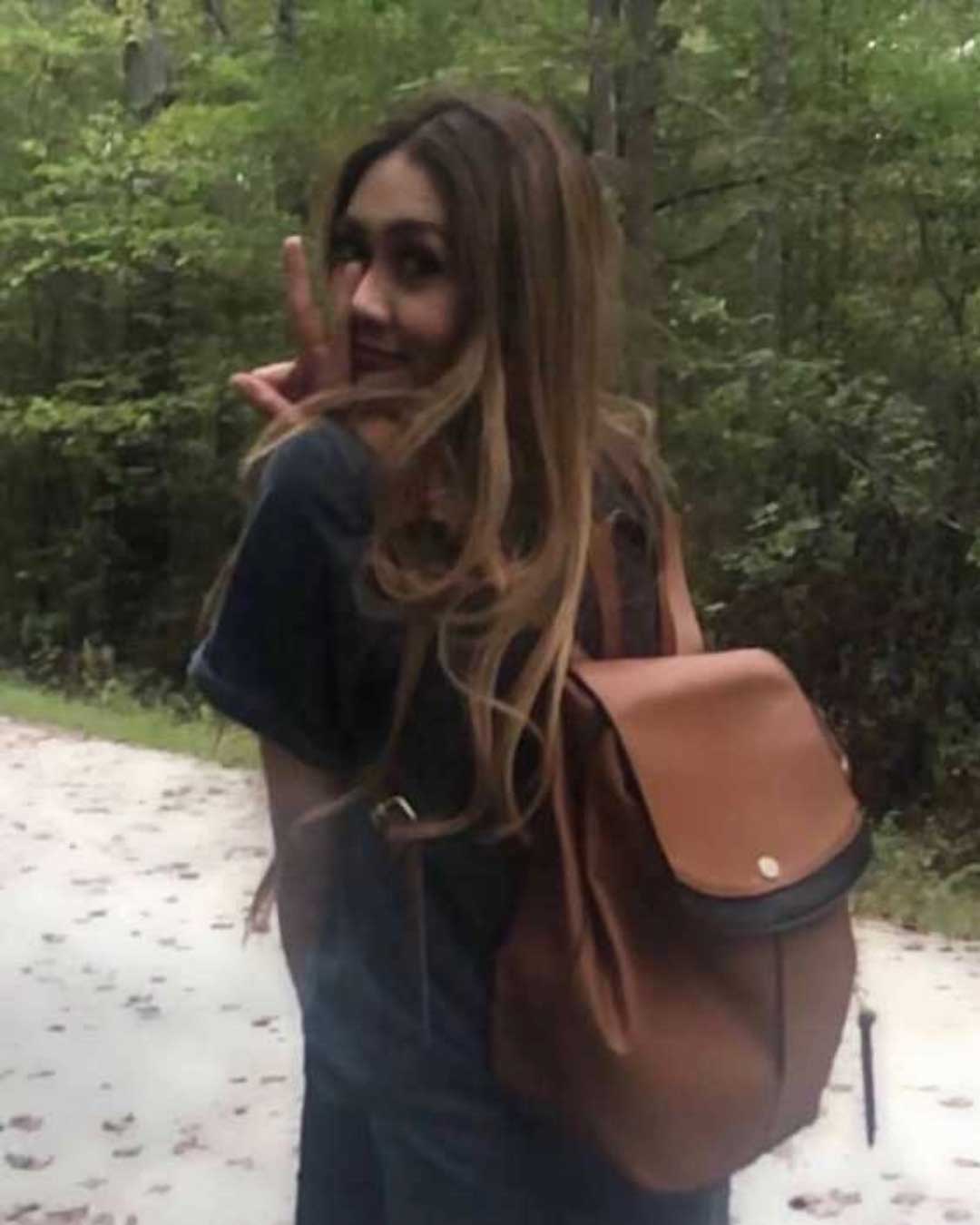 A person with long hair stands on a forest path. They are wearing a dark shirt and carrying a brown backpack. The person is turned slightly backward, smiling, and making a peace sign with their hand. The path is surrounded by greenery and trees.