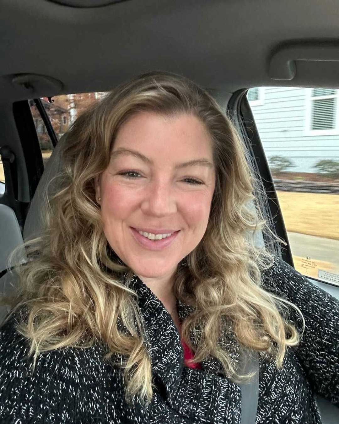 A woman with long, wavy blonde hair is sitting in a car and smiling at the camera. She is wearing a black patterned sweater and a red shirt. The background shows a residential street and a house with white siding.