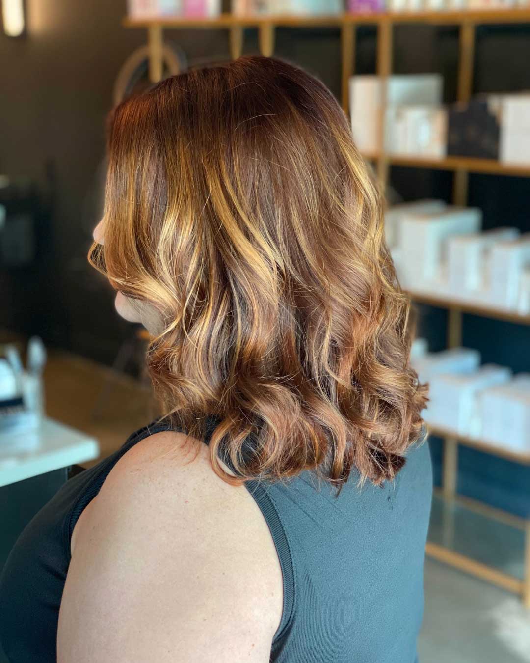 A person with shoulder-length, wavy brown hair featuring lighter highlights is seen from the side. They are in a well-lit room with shelves in the background holding various boxes and items. The person's hair appears freshly styled.