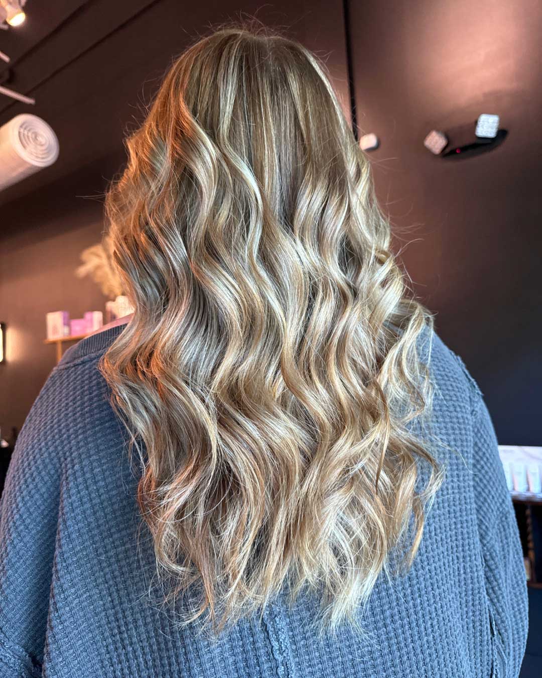 The image shows the back view of a person with long, wavy blonde hair cascading down their back. The hair has highlights and lowlights, creating a multidimensional effect. They are wearing a gray textured top. The background appears to be a dimly lit salon.