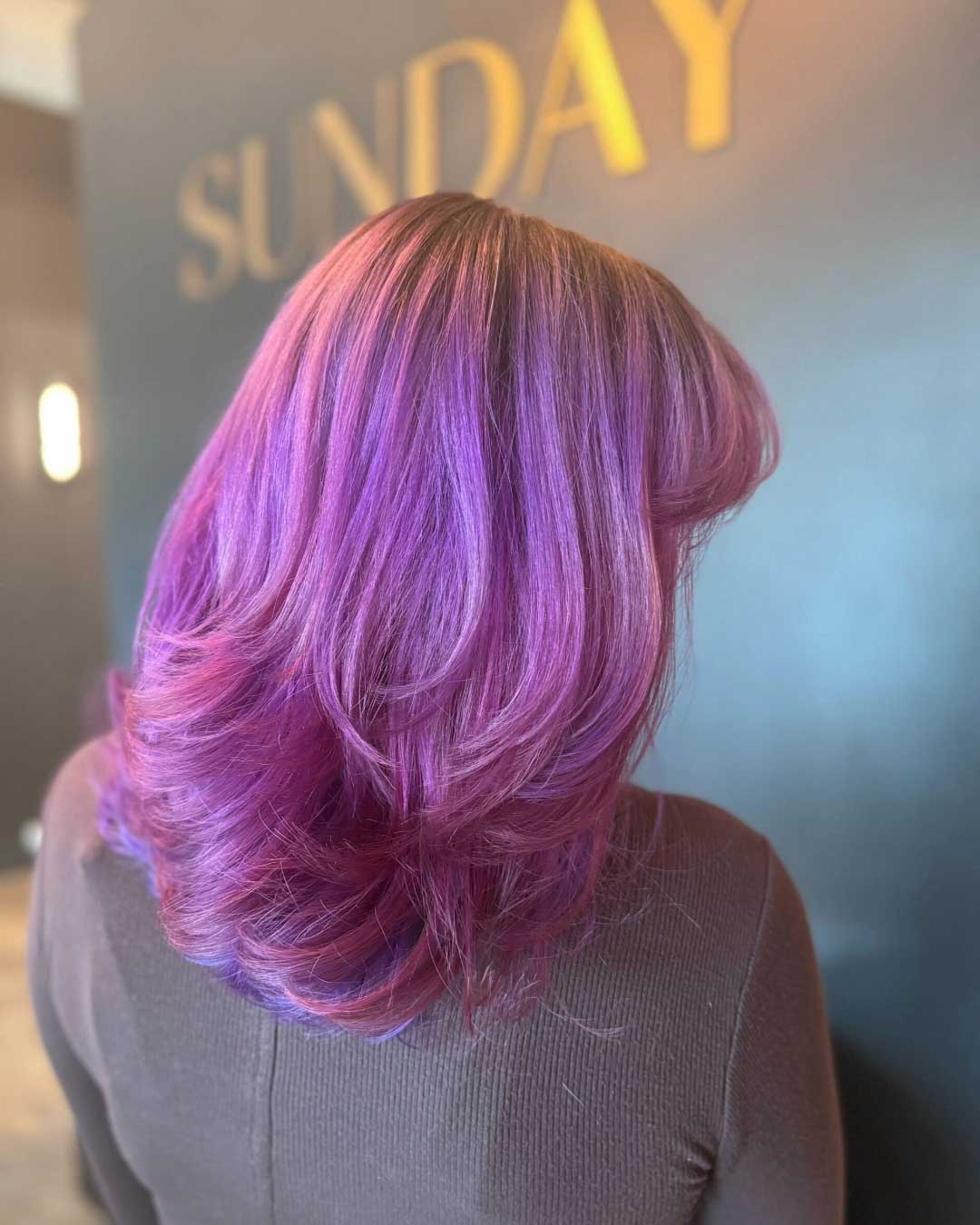 A person with shoulder-length, wavy hair dyed in a blend of vibrant purple and magenta hues stands in front of a dark wall with the word "SUNDAY" in large gold letters. They are wearing a dark top and the focus is on their hair from the back.