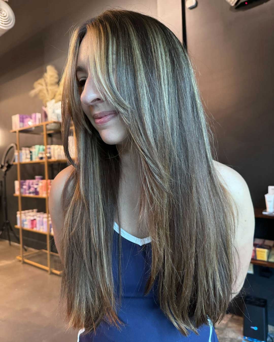 A young woman with long, straight hair featuring light brown and blonde highlights is standing in a room. She is smiling slightly and wearing a dark blue top. The background shows shelves with products and a dark-colored wall.