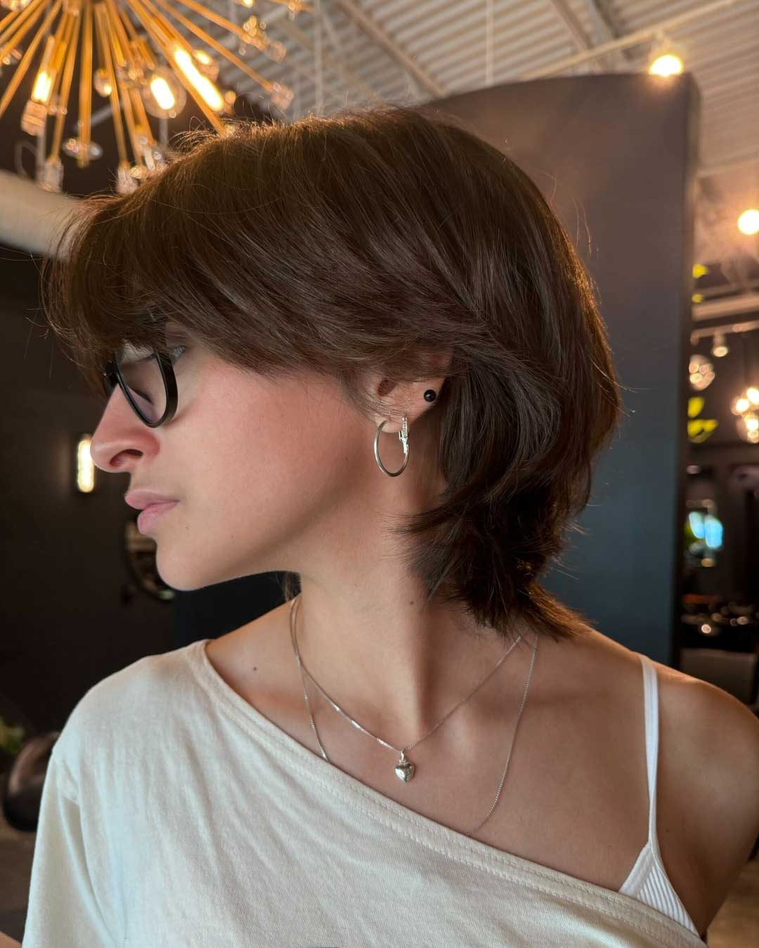 A person with short, layered brown hair and wearing glasses looks to the left. They are adorned with hoop earrings and a necklace. The individual is dressed in a light-colored off-the-shoulder top, and the background features modern, indoor lighting fixtures.