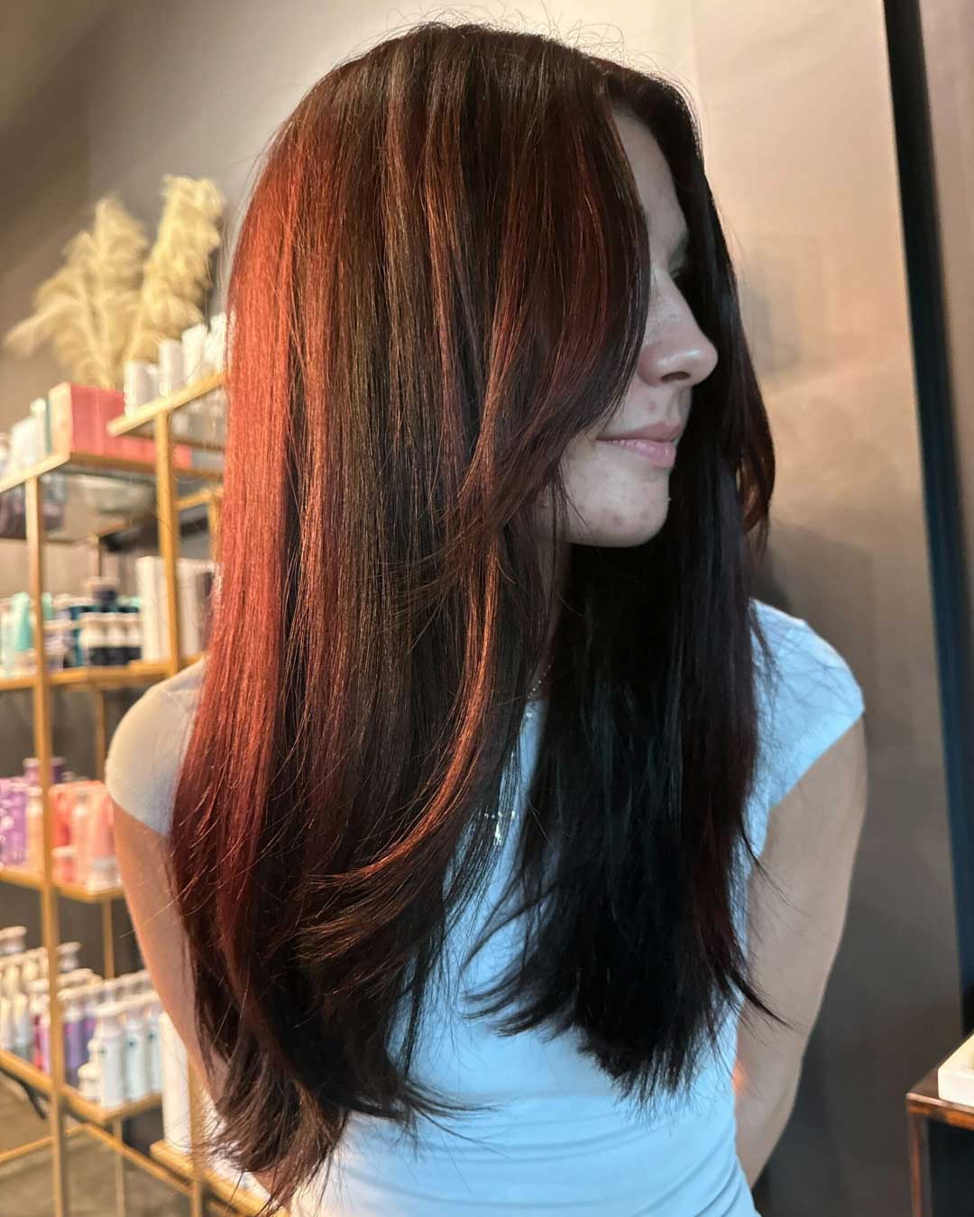 A woman with long, straight dark brown hair featuring subtle red highlights, wearing a white top, stands in profile. Behind her are salon shelves filled with various hair products. The lighting accentuates the hair's shine and color.