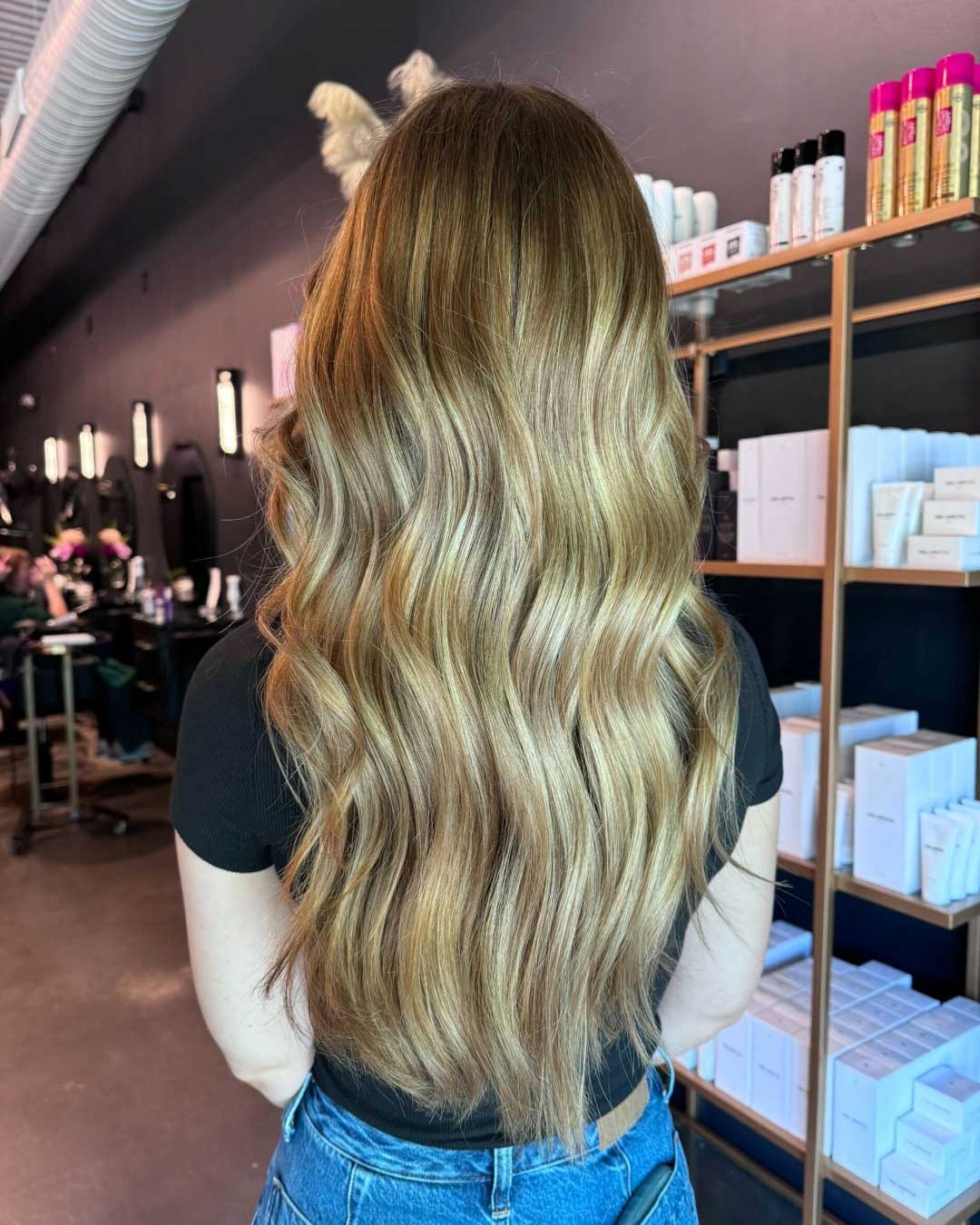 A person with long, wavy blonde hair stands in a hair salon, facing away from the camera. They are wearing a black shirt and jeans. The salon shelves in the background hold various hair care products, and other clients can be seen in the reflections of salon mirrors.