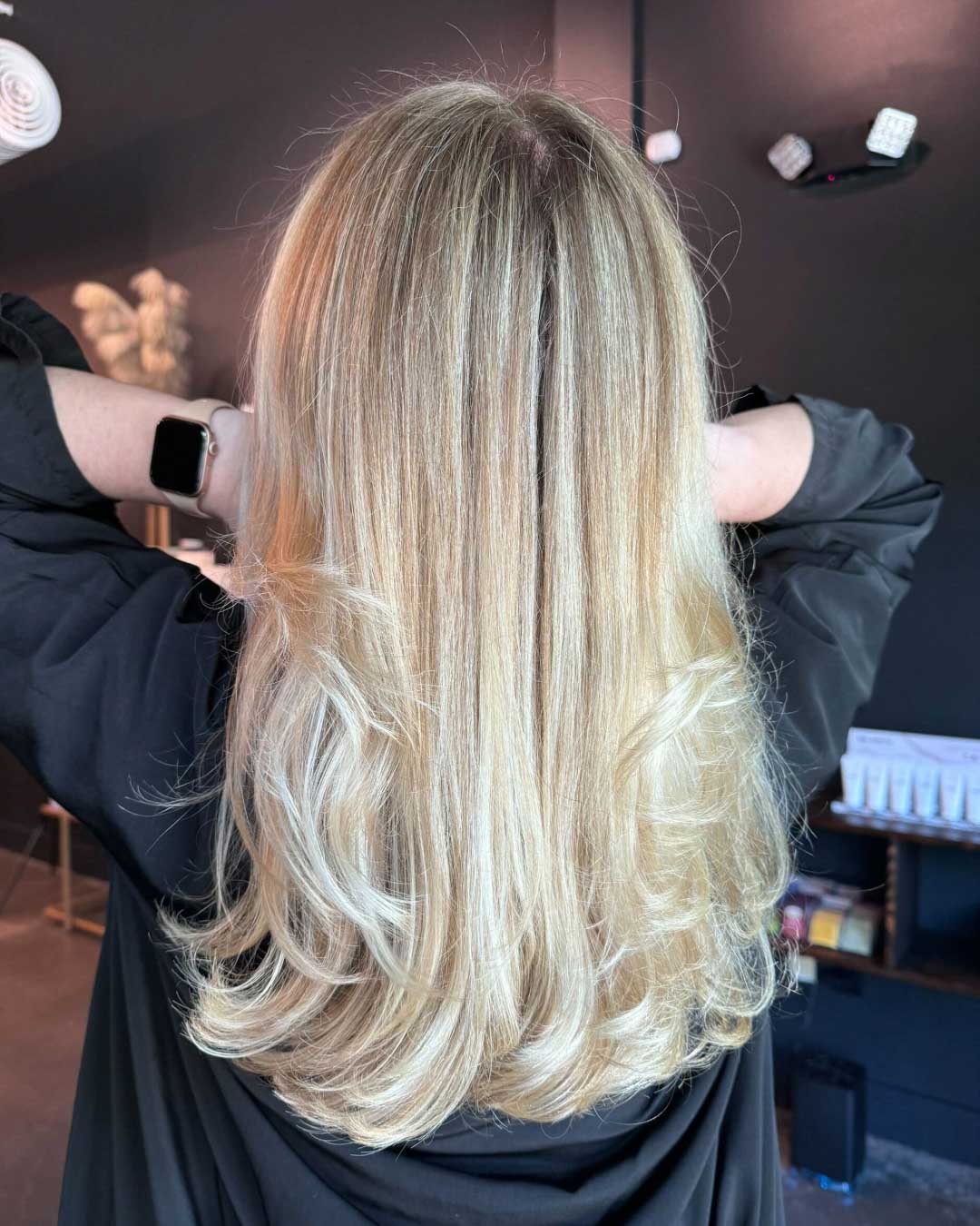 A person with long, straight blonde hair stands with their back to the camera, hands resting on their head. The hair has a smooth, shiny texture, and the setting appears to be a modern, dark-colored hair salon.