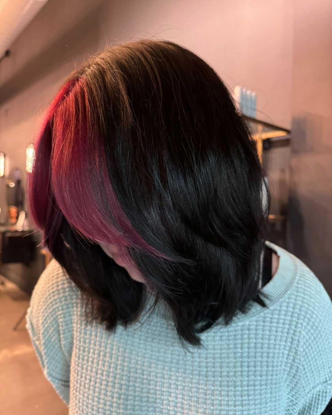 A person with shoulder-length, wavy black hair featuring a bright red streak on one side. They are indoors, wearing a light-colored textured sweater, and their head is tilted downwards, obscuring their face. The background is blurred with some shelves visible.