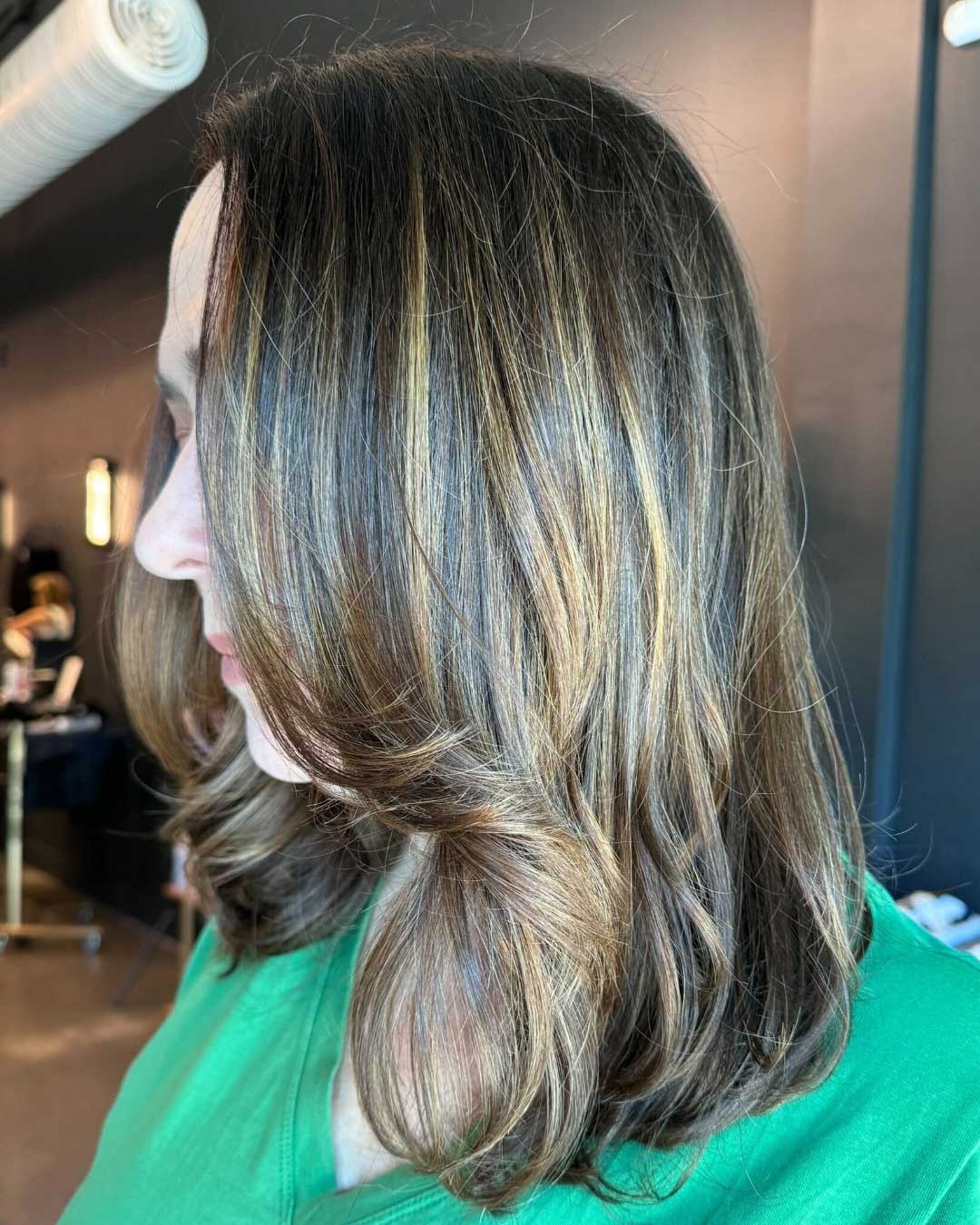 A person with shoulder-length brown hair highlighted with blonde streaks is shown in profile. The hair has soft waves and light curls, falling gently over their shoulder. The individual is wearing a green top, and the background is a dimly-lit indoor space.