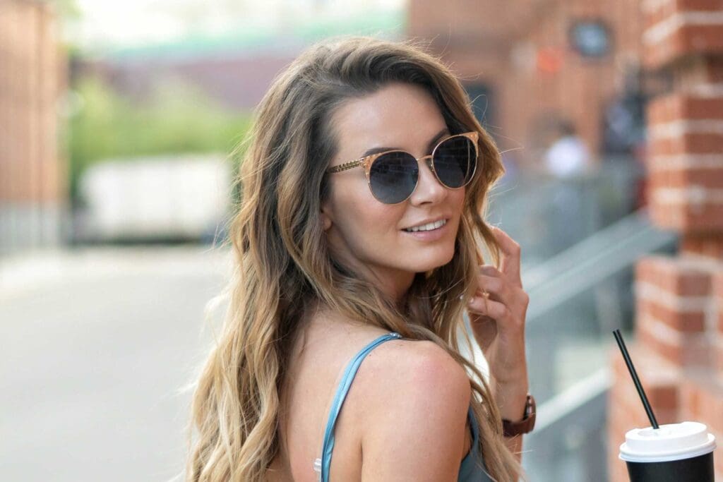 A woman with long, blonde balayage hair wearing sunglasses and a blue tank top smiles while turning her head. She stands outdoors in an urban setting, holding a cup with a straw. Brick buildings and a blurred background are visible.