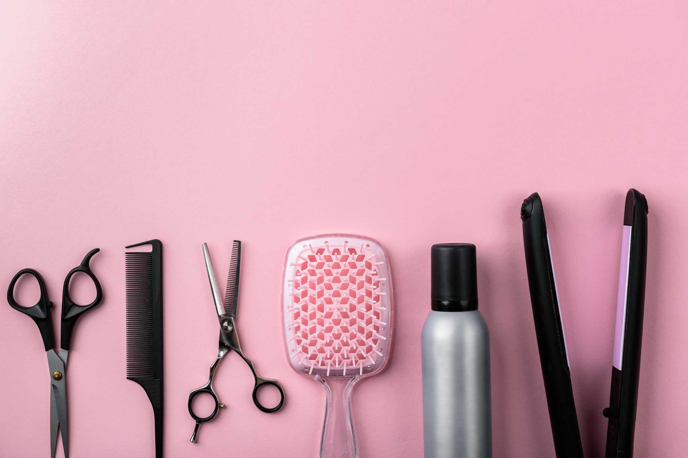 A flat lay of essential hair styling tools on a pink background, featuring precision scissors, a trusty comb, thinning shears, a chic pink hairbrush, an efficient spray bottle, and a sleek flat iron. Perfect for achieving salon-quality results right at home.