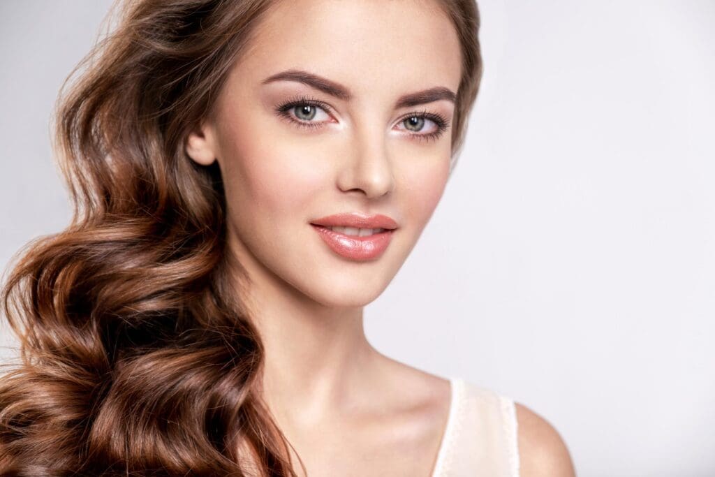 A woman with long, wavy brown hair featuring subtle balayage highlights and light makeup smiles softly at the camera. She is wearing a white top against a plain white background.